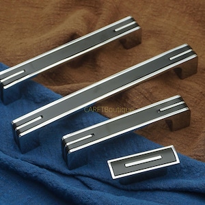 May include: A set of cabinet hardware with a modern design. The handles and knob feature a black center stripe with silver accents. The hardware is displayed on a blue and brown fabric background.