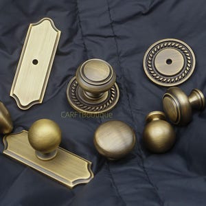 May include: An assortment of antique brass cabinet hardware, including knobs and backplates. The hardware features various designs, such as a round knob with a rope-like border and a rectangular backplate. The hardware is displayed on a dark, quilted surface.