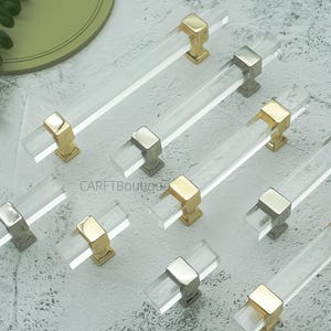 May include: Clear acrylic cabinet pulls with gold and silver-toned metal accents. The pulls are rectangular and have a modern design. Several pulls are arranged on a light grey surface. The text "CARFTBoutique" is visible.