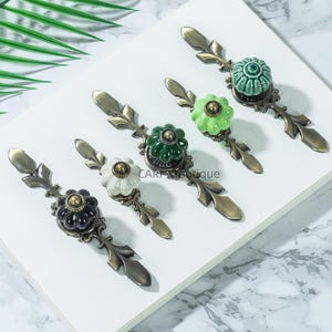 May include: A collection of decorative cabinet pulls with floral ceramic knobs in various colors: black, white, green, and teal. Each pull has an antique brass-colored metal base with a leaf design. The pulls are arranged on a white surface.