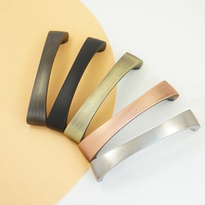 Può includere: Una collezione di cinque maniglie per mobili in varie finiture metalliche. Le maniglie sono curve e hanno una forma rettangolare. Le finiture includono bronzo, nero, ottone, rame e argento. Le maniglie sono disposte su uno sfondo bianco e beige.