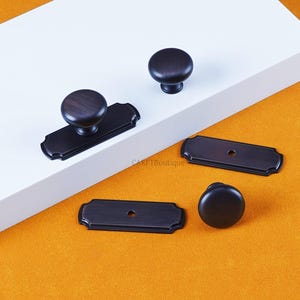 以下が含まれることがあります： 丸いノブと長方形のバックプレートを含む、濃い茶色のキャビネット金具のコレクション。 金具はオレンジ色の背景に対して白い表面に表示されます。 金具は木目調の外観をしています。