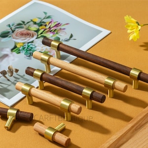 Może przedstawiać: Drewniane uchwyty do szafek z mosiężnymi detalami. Niektóre są ciemnobrązowe, inne z jasnego drewna. Uchwyty ułożone są na żółtej powierzchni, z kwiatowym nadrukiem w tle. Widoczny jest tekst "CARFTBoutique".