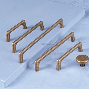 May include: A collection of antique brass cabinet hardware, including several bar pulls of varying lengths and a round knob. The hardware has a distressed finish and is arranged on a light gray surface. The brand name "CARFTBoutique" is visible.