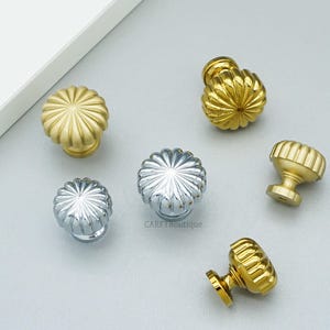 May include: A collection of decorative cabinet knobs in gold and silver finishes. The knobs feature a fluted, floral-like design. The hardware is displayed on a light gray surface. The knobs are suitable for furniture and home decor projects.