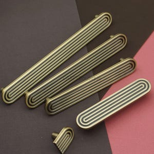 May include: A collection of antique brass cabinet hardware, including several elongated pulls and a small knob. The hardware features a grooved design with a dark accent, set against a brown and pink background.