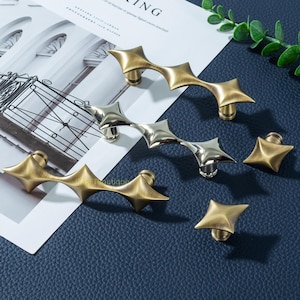 以下が含まれることがあります： 真鍮とシルバー仕上げの装飾キャビネットハードウェア。 ハードウェアには、星型のプルとノブが含まれています。 プルは複数の星のポイントで湾曲しています。 ノブは単一の星の形をしています。 ハードウェアは濃い青色の表面に表示されます。