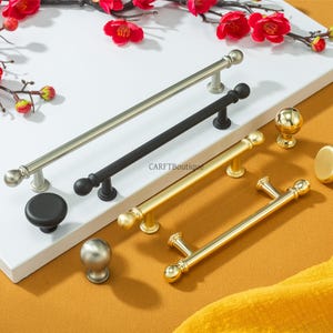 Może przedstawiać: Kolekcja uchwytów i gałek do szafek w kolorze srebrnym, czarnym i złotym. Uchwyty mają kształt prętów, a gałki są okrągłe. Okucia są prezentowane na białej powierzchni z czerwonymi kwiatami.