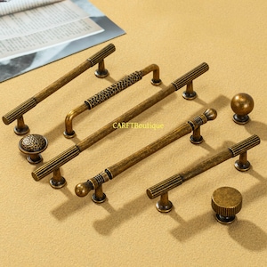 May include: An assortment of antique brass cabinet hardware, including handles and knobs. The handles vary in design, with ribbed and textured details. The knobs include a round, smooth knob and a knob with a textured surface. The hardware is displayed on a light tan surface.