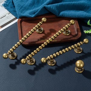 May include: Antique brass cabinet hardware set. Includes three beaded pull handles and one round knob. The hardware is displayed on a dark blue surface with a wooden tray and blue fabric in the background.