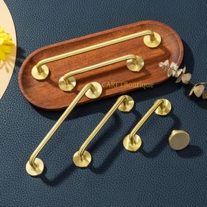 May include: A collection of brass-colored cabinet hardware, including handles and a knob, arranged on a wooden tray. The hardware has a modern, minimalist design with rounded edges and circular mounting plates. The tray sits on a dark blue surface.