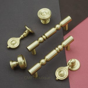 May include: An assortment of gold-toned cabinet hardware, including knobs and pulls. The hardware features a mix of cylindrical and spherical shapes, with a matte finish. The pieces are arranged on a two-tone background of gray and pink.