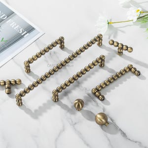 May include: An assortment of antique brass cabinet hardware, including handles and knobs. The handles are long and straight, adorned with a series of small, round beads. The knobs are spherical. The hardware is arranged on a white marble surface.