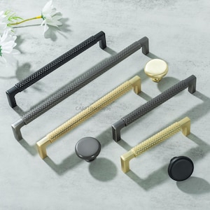 Puede incluir: Una variedad de tiradores y pomos para armarios en negro, gris y dorado. Los tiradores presentan un diseño texturizado con patrón de diamantes. Los pomos son redondos y vienen en varios acabados, ofreciendo opciones de diseño.