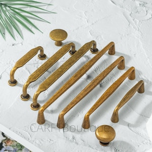 May include: An assortment of antique brass cabinet hardware, including knobs and various sizes of pull handles. The hardware has a distressed finish, giving it a vintage appearance. The hardware is arranged on a white surface.