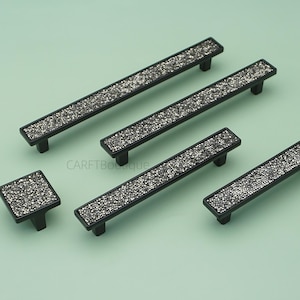 Może przedstawiać: Zestaw czarnych okuć do szafek, w tym uchwyty i gałka, z błyszczącą, kryształową wstawką. Okucia są pokazane na jasnym zielonym tle, oferując nowoczesną i efektowną estetykę.