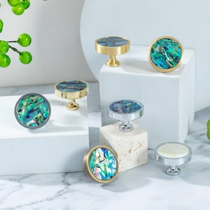 May include: A collection of decorative cabinet knobs with iridescent abalone shell inlays. The knobs feature various metal finishes, including gold, silver, and gunmetal. The knobs are round with a pedestal base, perfect for adding a touch of elegance to furniture.