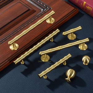 Puede incluir: Una colección de herrajes dorados para gabinetes con estilo de bambú, que incluyen manijas y perillas. El hardware se muestra sobre una superficie de madera oscura y una superficie azul similar al cuero, mostrando varios tamaños y diseños para la decoración del hogar.
