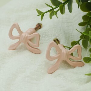 May include: Two pale pink bow-shaped cabinet knobs with brass-coloured screws. The knobs are made of a smooth material and are arranged on a white surface with green foliage in the background. The text "CARETBoutique" is visible.