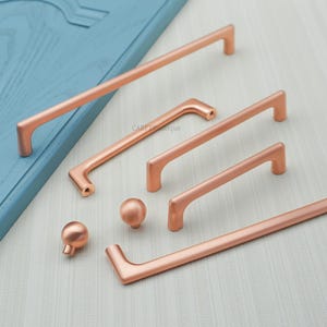 May include: A collection of copper-colored cabinet hardware, including various sizes of bar pulls and round knobs. The hardware is displayed against a light blue surface, showcasing the different designs and finishes available for kitchen or bathroom cabinetry.