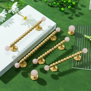 Op de afbeelding: Een verzameling messing kastgrepen en knoppen met roze edelsteenaccenten. De grepen hebben een getextureerd, geribbeld ontwerp en zijn afgewerkt met een roze bol. De knoppen hebben ook een roze bol. De hardware wordt gepresenteerd op een groene ondergrond.