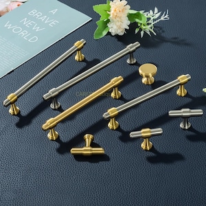May include: An assortment of cabinet hardware, including handles and knobs, in gold and silver tones. The hardware is displayed on a dark blue surface, with a magazine that reads "A Brave New World" in the background.