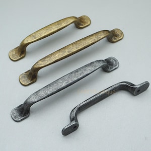 May include: Four antique-style cabinet pulls in varying shades of bronze and gray. The pulls have a curved handle design with decorative end pieces. The hardware is made of metal and has a distressed finish, suggesting an aged appearance.