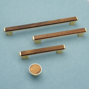 Könnte beinhalten: Ein Set aus Holzschrankbeschlägen mit modernem Design. Das Set enthält drei rechteckige Griffe und einen runden Knopf. Die Griffe haben eine dunkle Holzmitte mit goldfarbenen Metallenden. Der Knopf hat eine passende Holzmitte und einen goldenen Sockel.