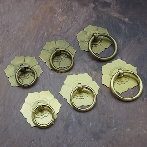 May include: Six antique brass drawer pulls. Each pull features a floral base with a circular handle. The pulls are arranged on a mottled brown surface. The pulls are likely for furniture or cabinetry.
