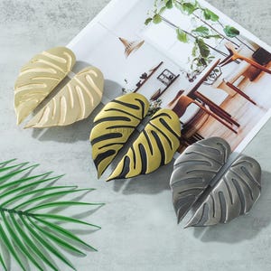 May include: Decorative leaf-shaped cabinet pulls in gold, black and gold, and silver finishes. The pulls are arranged on a light gray surface with a magazine and a green leaf in the background. The pulls have a textured design.