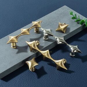 Op de afbeelding: Een verzameling decoratieve meubelbeslag, waaronder stervormige knoppen en handgrepen. De handgrepen hebben een uniek, geometrisch ontwerp met een messing afwerking, terwijl één handgreep en een knop een zilveren afwerking hebben. Gepresenteerd op een grijze ondergrond tegen een blauwe achtergrond.