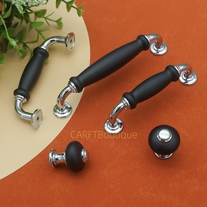 Peut inclure: Ensemble de poignées et de boutons de meubles noirs et chromés. Les poignées ont un design incurvé avec un centre noir et des accents chromés. Les boutons sont ronds avec un dessus noir et une base chromée. Le texte "CARETBoutique" est visible.