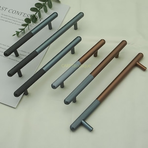 Peut inclure: Six poignées de meubles de différentes couleurs : gris foncé, bleu-gris et bronze. Chaque poignée a une section centrale côtelée et une poignée lisse et arrondie. Les poignées sont disposées sur une surface claire.
