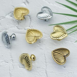 May include: Heart-shaped cabinet knobs in gold and silver finishes. The knobs have a textured, fan-like design. The hardware is shown on a white textured surface. Decorative cabinet pulls for furniture.