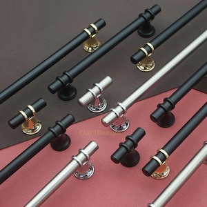 Peut inclure: Divers modèles de poignées et boutons de meubles, noirs, argentés et dorés. Les poignées sont cylindriques avec des anneaux décoratifs, et les boutons sont en forme de T. Présentation sur un fond marron et rouge.