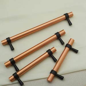 May include: Four copper-colored cabinet pulls with black accents. Each pull features a cylindrical bar with black end pieces and a black handle. The pulls are arranged diagonally on a light-colored surface.