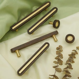 Peut inclure: Ensemble de poignées et boutons de meubles en bois. Les poignées sont rectangulaires, avec une finition en bois brun foncé et une bande centrale dorée. Les boutons sont ronds avec un centre doré. La quincaillerie a des montants dorés.