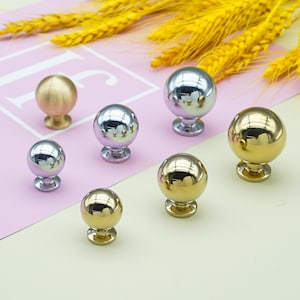 Może przedstawiać: Kolekcja błyszczących, kulistych gałek do szafek w odcieniach złota i srebra. Gałki mają klasyczny kulisty kształt z ozdobną podstawą. Tło przedstawia kłosy pszenicy oraz różową i białą powierzchnię.