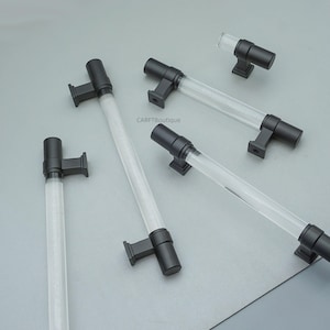 Może przedstawiać: Kilka uchwytów do szafek z przezroczystym, cylindrycznym środkiem i czarnym osprzętem. Uchwyty są ułożone na szarej powierzchni. Przezroczysty środek wydaje się być wykonany z akrylu lub szkła. Osprzęt jest kwadratowy i prostokątny.