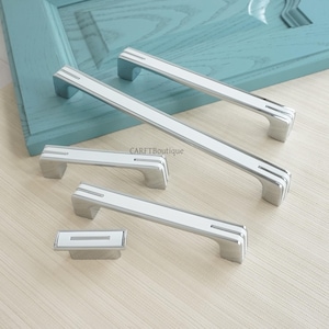 Könnte beinhalten: Eine Sammlung von silbernen und weißen Schrankgriffen und -knöpfen. Die Hardware hat eine rechteckige weiße Mitte mit silbernen Akzenten und ein gebogenes Griffdesign. Der Markenname "CARFTBoutique" ist sichtbar.