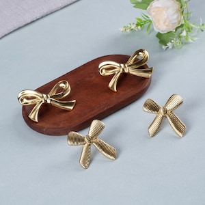 May include: Four gold-colored bow-shaped decorative hardware pieces. Two are on a dark brown wooden oval, and two are separate. The bows have a shiny, metallic finish and are arranged on a light blue surface.