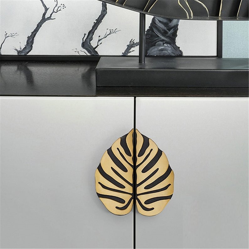 May include: A cabinet with a gold and black leaf-shaped handle. The cabinet doors are white, and the handle is in the shape of a tropical leaf. The background features a black shelf and a painting with black and white tree branches.