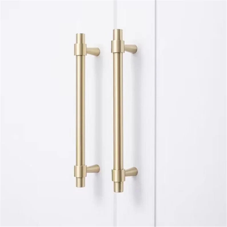 Peut inclure: Deux poign&eacute;es de meuble dor&eacute;es avec un design en T. Les poign&eacute;es sont mont&eacute;es sur une surface blanche. Chaque poign&eacute;e a un corps cylindrique avec une poign&eacute;e en forme de T &agrave; chaque extr&eacute;mit&eacute;. Le mat&eacute;riel est en m&eacute;tal.