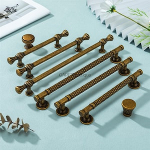 Peut inclure: Ensemble de quincaillerie de meubles en bronze antique, comprenant des boutons et des poignées de différentes tailles. La quincaillerie présente des détails décoratifs et une finition vieillie et chaleureuse. Les articles sont disposés sur une surface bleu clair.