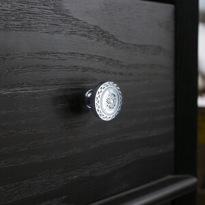 May include: Close-up of a silver drawer pull with an intricate circular design. The pull is mounted on a dark, textured wooden drawer front. The design features a central pattern surrounded by geometric shapes.