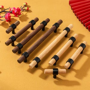 Może przedstawiać: Kolekcja drewnianych uchwytów do szafek w różnych rozmiarach i odcieniach drewna, w tym ciemnobrązowym i jasnym drewnie. Każdy uchwyt posiada czarny metalowy akcent i czarną podstawę. Uchwyty są ułożone na żółtej powierzchni.