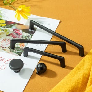 May include: A selection of black cabinet hardware, including three pull handles of varying lengths and two knobs. The handles are rectangular with curved ends. The knobs are a round, flat-topped knob and a spherical knob. The background is a warm yellow.