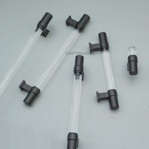 K&ouml;nnte beinhalten: Mehrere transparente Acrylr&ouml;hren mit schwarzen Beschl&auml;gen sind auf einer hellgrauen Oberfl&auml;che angeordnet. Die R&ouml;hren haben eine zylindrische Form und sind mit schwarzen rechteckigen Halterungen verbunden. Eine kleinere R&ouml;hre mit schwarzen Beschl&auml;gen ist ebenfalls vorhanden. Der Text "CARFTBoutique" ist sichtbar.