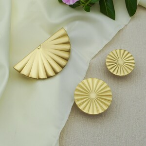May include: A set of gold-coloured cabinet hardware, comprising two round knobs and a half-circle pull. The hardware has a fan-like design with radiating lines. The items are displayed on a cream-coloured fabric.