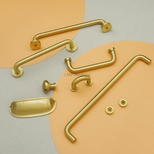 May include: A selection of brass-toned cabinet hardware, including handles, knobs, and a small tray. The hardware has a smooth, matte finish and a simple, modern design. The pieces are arranged on a peach and grey background.